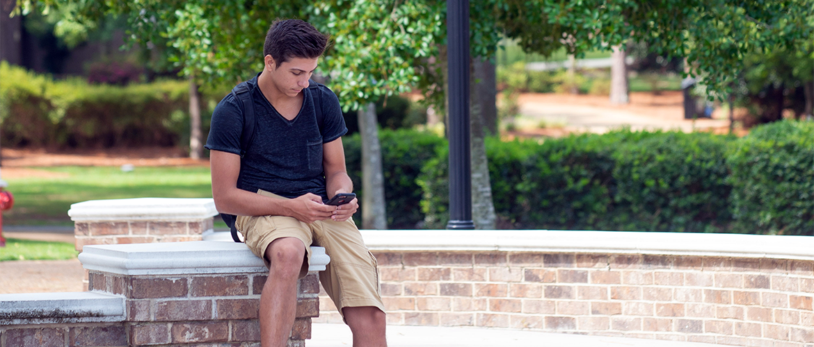Student sitting on a brick pillar while scrolling on phone