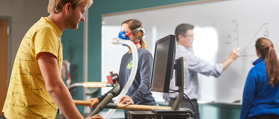 Student on a treadmill in the foreground, student on a breathing machine in the middle, professor and student writing on a whiteboard in the background